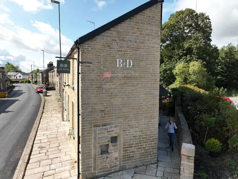 Bollington Dental Practice from above: a stone cottage on a village street, the practice name on its gable end and a hanging sign over the pavement, with a patient walking up the garden path